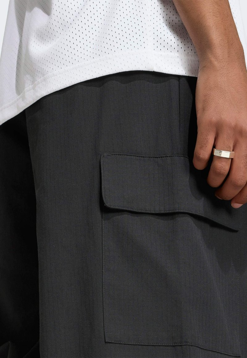 Main d'homme avec une bague en argent reposant près de la poche à rabat d'un pantalon cargo noir, portant une chemise blanche texturée.