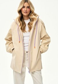 Kobieta o długich blond włosach, ubrana w beżową dużą kurtkę z kapturem z fioletowymi troczkami, białą koszulę i jasne spodnie.