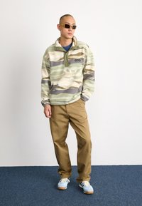 Pullover in fleece mimetico verde, beige e grigio con zip a quarto e tasca con patta; abbinato a pantaloni khaki e sneakers azzurri chiari.