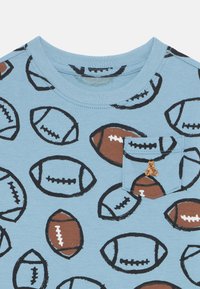 Hellblaues T-Shirt mit kurzen Ärmeln und einem sich wiederholenden Fußballmuster in Braun und Schwarz. Verfügt über eine kleine Tasche mit einer Bärenstickerei.