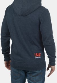 Mikina s kapucí v tmavě modré tkanině s žebrovanými manžetami a lemem. Na dolní části zad je červenobílé logo. Volný střih.