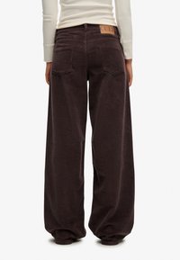 Pantalones de pana de pierna ancha en marrón oscuro. Textura acanalada, grandes bolsillos traseros y un parche de cuero color beige en la cintura.