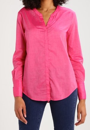 Žena oblečená v jasně růžové blůze s dlouhými rukávy a výstřihem do V, v kombinaci s tmavě modrými džínami, stojící před jednobarevným bílým pozadím.