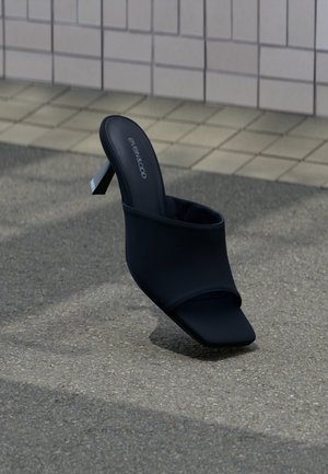 Fekete, nyitott orrú papucs vastag sarokkal és négyzet alakú orral, az "EVEN&ODD" márkajelzéssel, szürke járdán egy csempézett fal közelében elhelyezve.