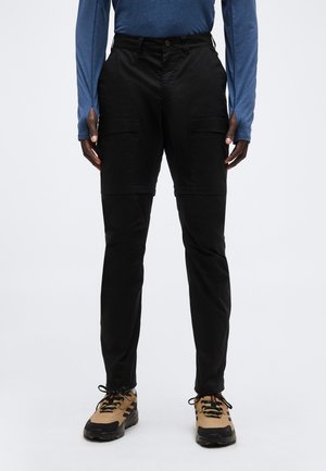 Άτομο που φοράει μαύρο παντελόνι cargo με τσέπες με καπάκι, μπλε μακρυμάνικο πουκάμισο και μπεζ παπούτσια πεζοπορίας, στέκεται μπροστά σε απλό φόντο.