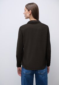 Camisa de manga larga marrón oscuro con cuello puntiagudo, que presenta puños abotonados y un corte relajado. Combinada con jeans azules.