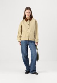 Giacca beige con bottoni e colletto marrone, due tasche frontali, abbinata a jeans blu a gamba larga e una borsa verde a spalla.