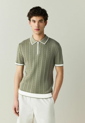 Göndör hajú fiatal férfi, zöld, kábel mintás rövid ujjú galléros pólót visel fehér szegéllyel és fehér nadrágot, egy egyszerű háttér előtt áll.