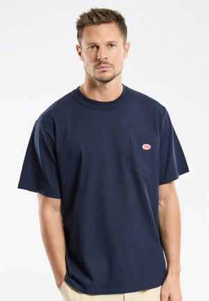 Man wearing navy blue short-sleeve T-shirt with chest pocket and small red logo, standing against a plain white background.