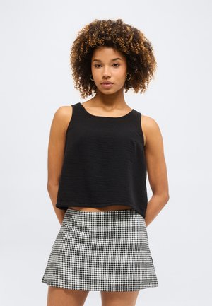 Γυναίκα με σγουρά μαλλιά, που φορά χρυσούς κρίκους στ' αυτιά, μαύρο αμάνικο τοπ και ασπρόμαυρη καρό φούστα, στέκεται με τα χέρια πίσω από την πλάτη.