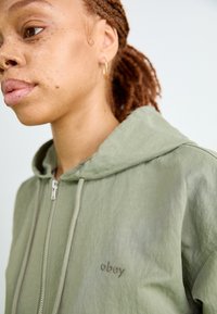 Svetlozeleno zip-up kapuča z teksturiranim zaključkom, kapuca s vrvico in vezenim logotipom "obey". Ima srebrno zadrgo in mehak material.