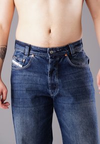 Denim jeans in dark blue with a faded texture, five pockets, copper rivets, and a zip fly. No visible logos or distinct patterns.