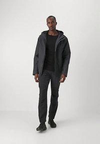 Man som bär en svart huva jacka, svart skjorta, svarta byxor och svarta skor, står mot en enfärgad ljus bakgrund.