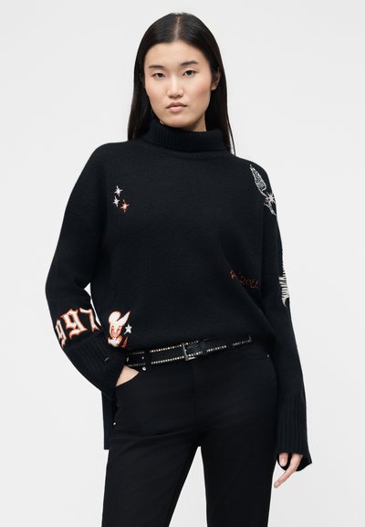 Femme aux longs cheveux noirs portant un pull à col roulé noir avec des patchs blancs et orange, un pantalon noir et une ceinture cloutée, debout avec une main dans la poche.