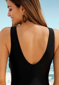 Maillot de bain noir une pièce avec un design dos en V profond, panneaux latéraux texturés et larges bretelles. Porté avec de grandes boucles d'oreilles marron sur fond de plage.