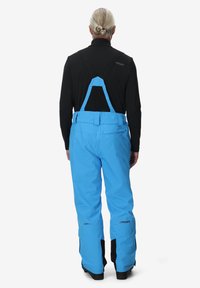 Persoon met grijs haar in een knot draagt een zwart langemouwen top en felblauwe skibroek met zwarte bretels, staande met de rug naar ons gekeerd.