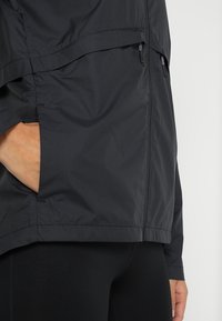 Veste légère noire avec un extérieur lisse, dotée d'une poche zippée sur le côté et d'une coupe décontractée. Le tissu semble résistant à l'eau.