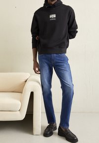 Svart huvtröja med logotyp, blå jeans och svarta skor. Modellen står bredvid en ljus soffa och visar upp avslappnad klädsel.