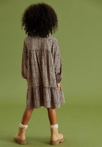 Pitkähihaista mekkoa beigen värisessä kankaassa, jossa on ruskea leopardikuosi, kerroksellinen muotoilu ja ryppytysdetaljit. Asusteena on vaaleanruskeat nilkkurit ja kermaiset sukat.