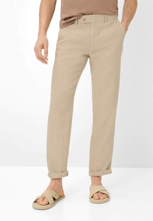 Mand iført beige bukser med opkrogede ben, lysebrun t-shirt og beige sandaler med krydsremme, stående med den ene hånd i lommen.