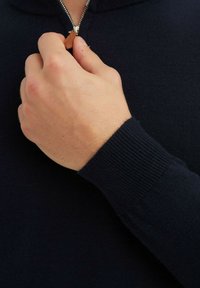 Pull zip bleu marine avec des poignets côtelés. Gros plan d'une main tenant la fermeture éclair métallique, mettant en valeur la texture tricotée et la languette en cuir.