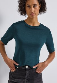 Haut court teal à manches courtes en tissu doux avec un col rond et des manches légèrement bouffantes, associé à un jean noir, les mains sur les hanches.