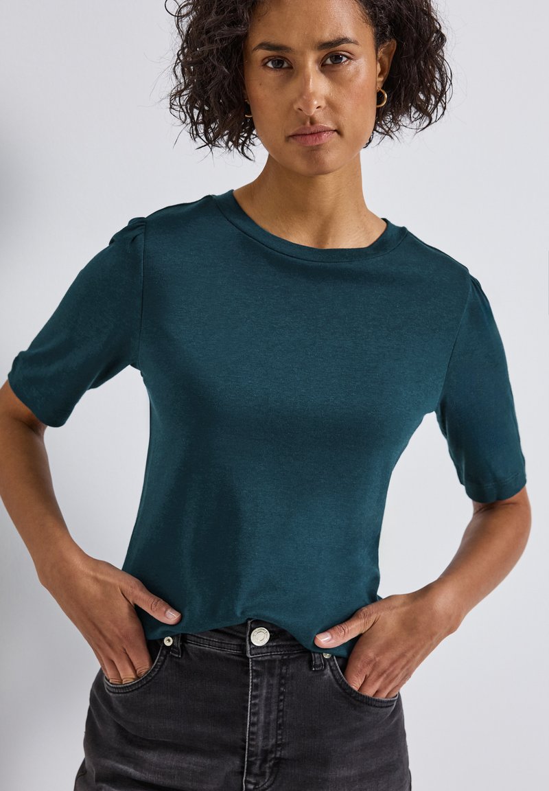 Haut court teal à manches courtes en tissu doux avec un col rond et des manches légèrement bouffantes, associé à un jean noir, les mains sur les hanches.