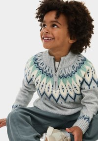 Maglione grigio in lana con colletto zip che presenta un motivo geometrico in verde, blu e bianco. Abbinato a pantaloni grigi larghi e scarpe chiare.