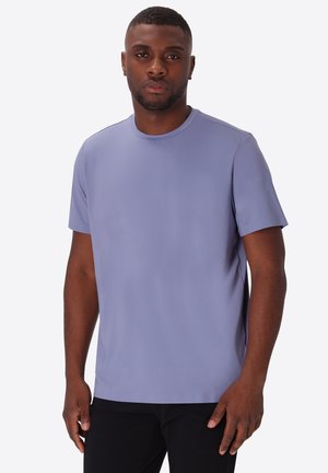 Uomo in piedi che indossa una maglietta tinta unita viola chiaro a maniche corte e pantaloni neri su uno sfondo bianco uniforme.