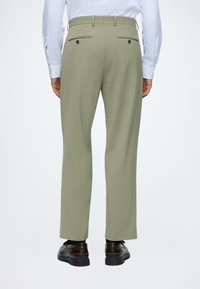 Homme portant un pantalon de costume olive clair, une chemise blanche à manches longues et des chaussures de ville marron foncé, vu de dos sur un fond uni.