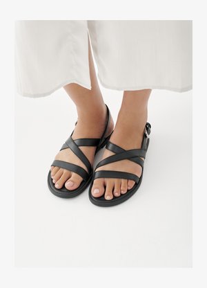 Voeten in zwarte sandalen met bandjes en enkelgespen, gecombineerd met een wit kledingstuk dat net boven de enkels eindigt, op een witte achtergrond.