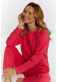 Różowy zestaw bawełnianych bluz i spodni z długim rękawem, ściągaczami oraz subtelnym detalem z logo. Modelka siedzi, uśmiechając się, w białych trampkach.