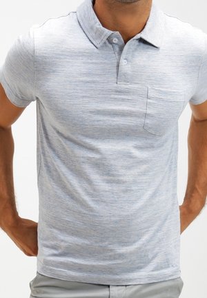 Man wearing a light grey short-sleeve polo shirt with a buttoned collar and a chest pocket, standing with hands behind his back.