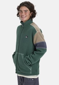 Grüne Fleecejacke mit sandfarbenen und marineblauen Seitenpaneelen. Sie hat einen hohen Kragen, einen Reißverschluss an der Vorderseite und zwei seitliche Taschen. Strukturierter Stoff mit weißen Nähten.