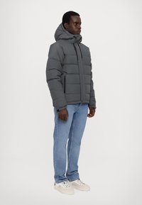 Veste grise matelassée avec capuche, dotée d'une fermeture éclair à l'avant, de poches latérales et d'une coupe carrée, associée à un jean bleu clair et à des baskets blanches.
