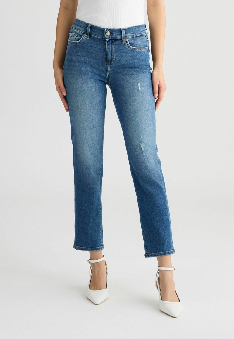 Femme portant un jean bleu taille mi-haute légèrement usé, un haut blanc sans manches et des escarpins blancs pointus, debout.