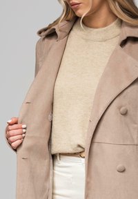Beżowy dwurzędowy płaszcz z zaokrąglonymi guzikami i miękką, teksturowaną tkaniną, noszony na jasnobeżowym swetrze i białych spodniach.