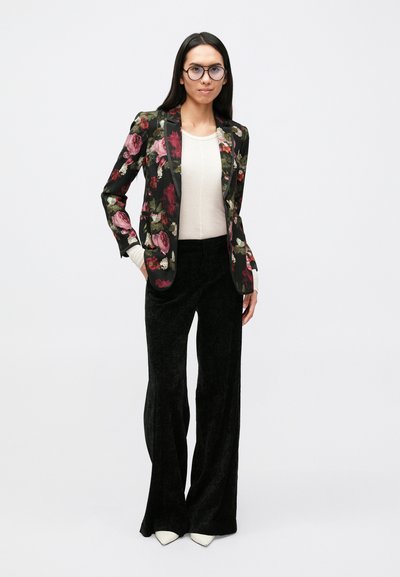 Blazer noir à motif floral, cintré avec des revers crantés, associé à un pantalon large noir texturé et des chaussures blanches à bout pointu.