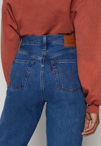Höga blå denimjeans med en urblekt textur, orange sömmar och två bakfickor, med en brun läderetikett på midjebandet.
