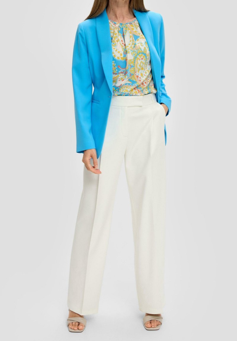 Femme portant un blazer bleu vif, un chemisier à motifs, un pantalon blanc taille haute à jambes larges et des talons ouverts beige, une main dans la poche.