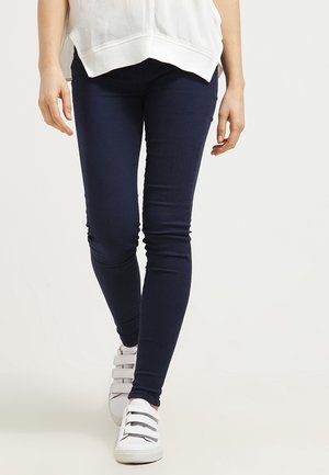 Person iført marineblå skinny bukser, hvide sneakers med velcrolukninger og en løs hvid top, der går fremad.