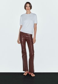 Un modello si staglia su uno sfondo neutro, indossando una maglietta grigia chiara, pantaloni in pelle marrone e sandali rossi, mentre assume una posa informale.