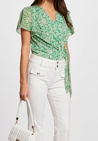 Top avvolgente floreale verde con maniche a campana corte, abbinato a pantaloni bianchi a vita alta con dettagli in bottone dorato e tasche. Tiene una borsa bianca.