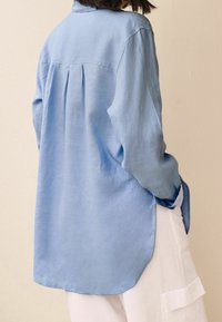 Chemise en lin bleu clair à coupe décontractée, dotée d'un pli au dos, de manches longues et d'un ourlet courbé, associée à un pantalon blanc.