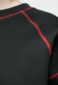 Fekete pulóver kerek nyakkivágással, kontrasztos piros varrással a varratoknál és a vállakon. Puha tapintású, pamutkeverék anyag.