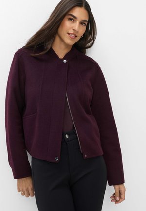 Donna con lunghi capelli scuri che indossa una giacca viola scuro con zip e pantaloni neri, in piedi su uno sfondo bianco.