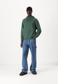 Grønn hettegenser med kengurulomme, paret med løse blå jeans med sidelommer. Svart caps og mørke joggesko fullfører antrekket.
