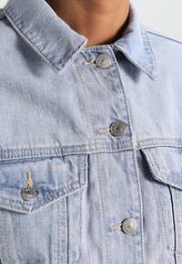 Světlemodrá džínová bunda s límečkem košile, dvěma náprsními kapsami a stříbrnými knoflíky s rytými detaily. Texturovaná tkanina s viditelným šitím.