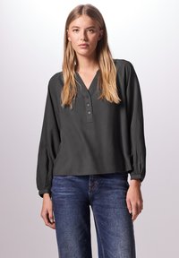 Blusa grigia scura a maniche lunghe con scollatura a V, bottoni sul davanti e polsini elastici, abbinata a jeans blu con micro pois. Tessuto dalla consistenza liscia.