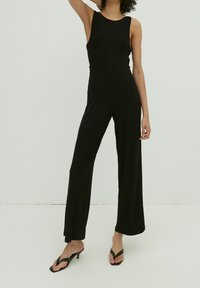 Mono negro de canalé con un diseño sin mangas, pantalones de pierna ancha y cintura ajustada. La modelo lleva sandalias negras de tacón. Sencillo, color sólido.